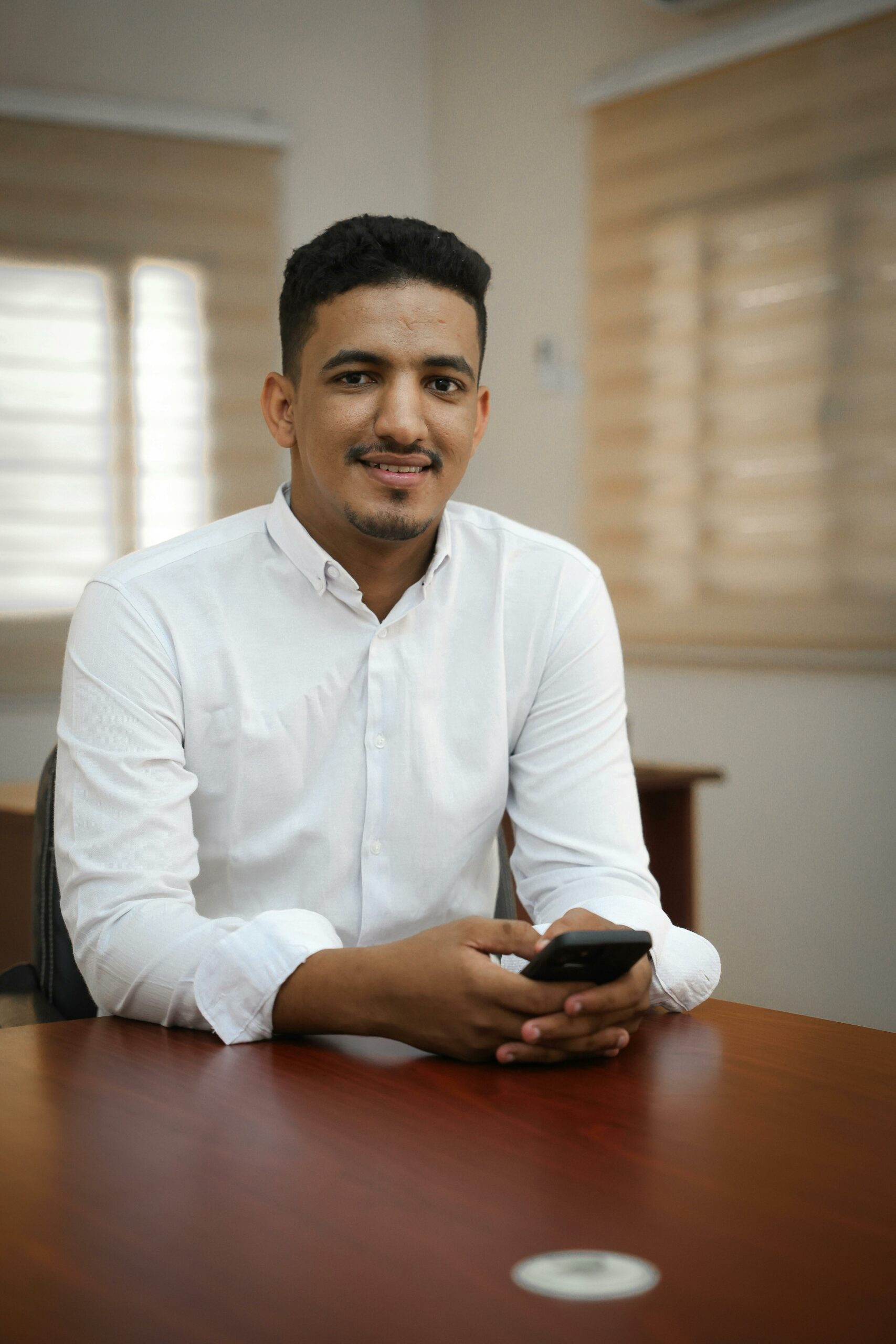 Confident young man in office setting with smartphone, smiling at camera.